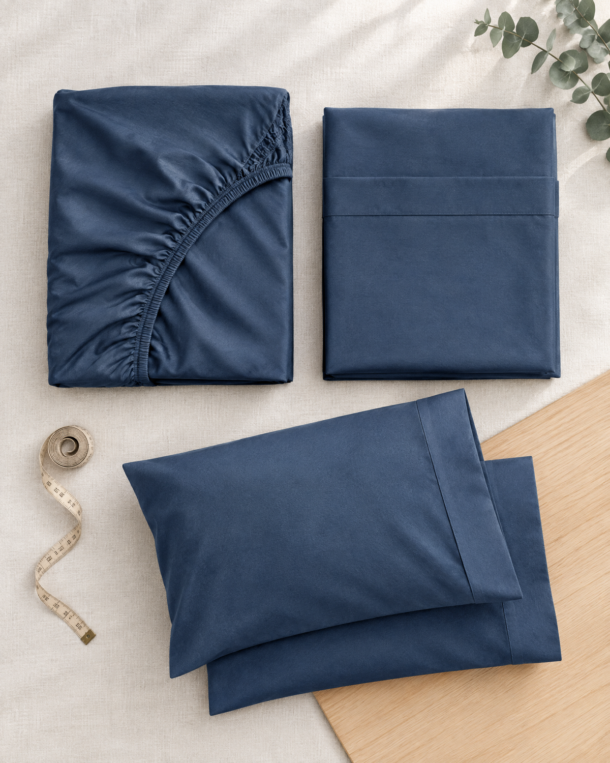 Overhead view of deep slate-blue bamboo viscose bedding showing a fitted sheet with elasticated edges, a folded flat sheet, matching pillowcases, and a measuring tape.