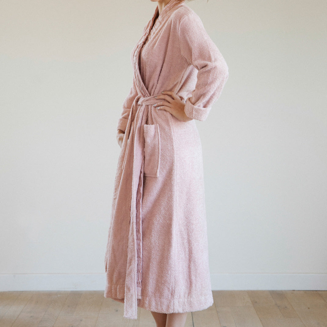 Woman wearing a pink robe standing against a white wall.